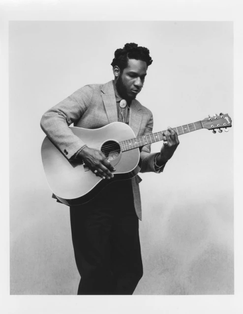 Black and white portrait of African American male playing an acoustic guitar, wearing a blazer and bolo tie, against a neutral background.