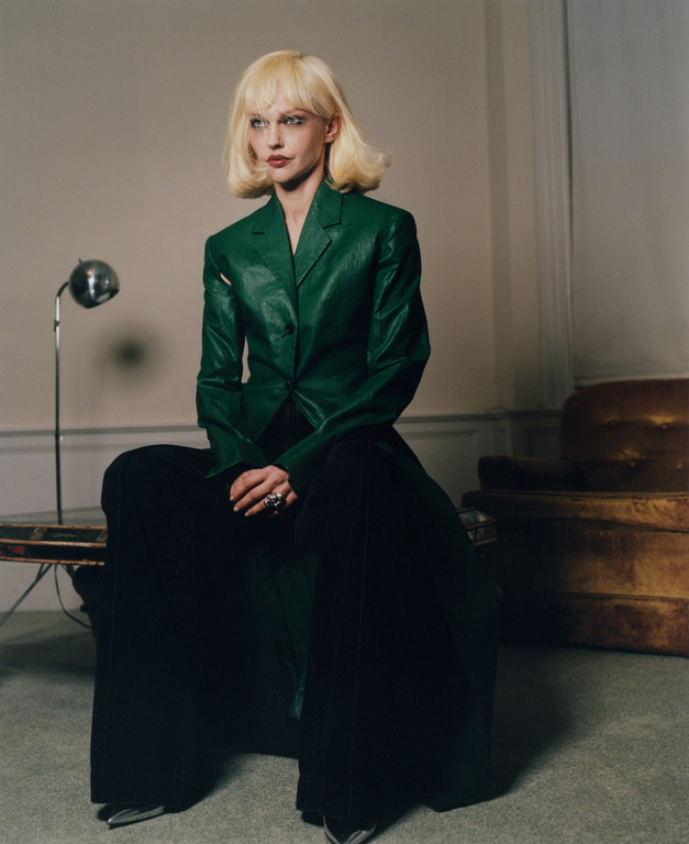 Adult woman in a green jacket and black pants sits on a table in an indoor setting with neutral walls and a vintage couch in the background.