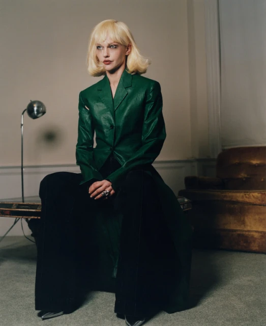 Adult woman in a green jacket and black pants sits on a table in an indoor setting with neutral walls and a vintage couch in the background.