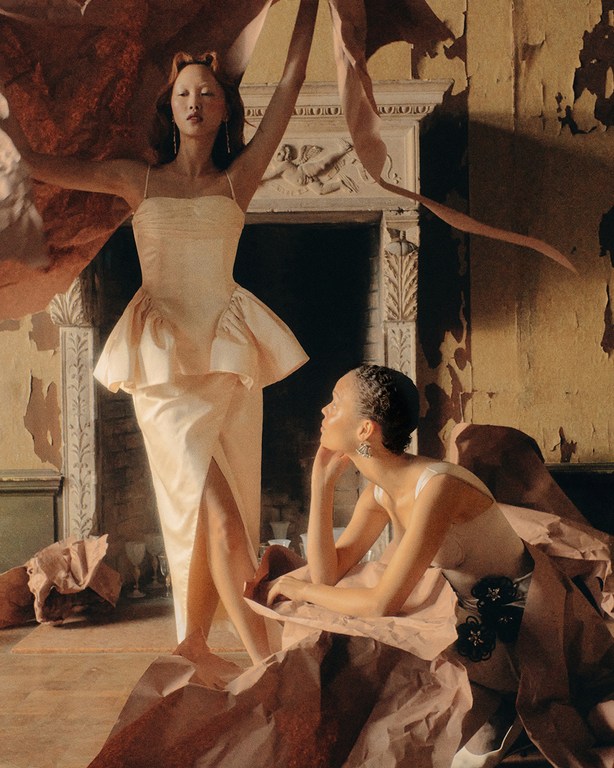 Two women in formal dresses pose in an ornate room. One woman stands with arms raised; the other sits with her head resting on her hand.
