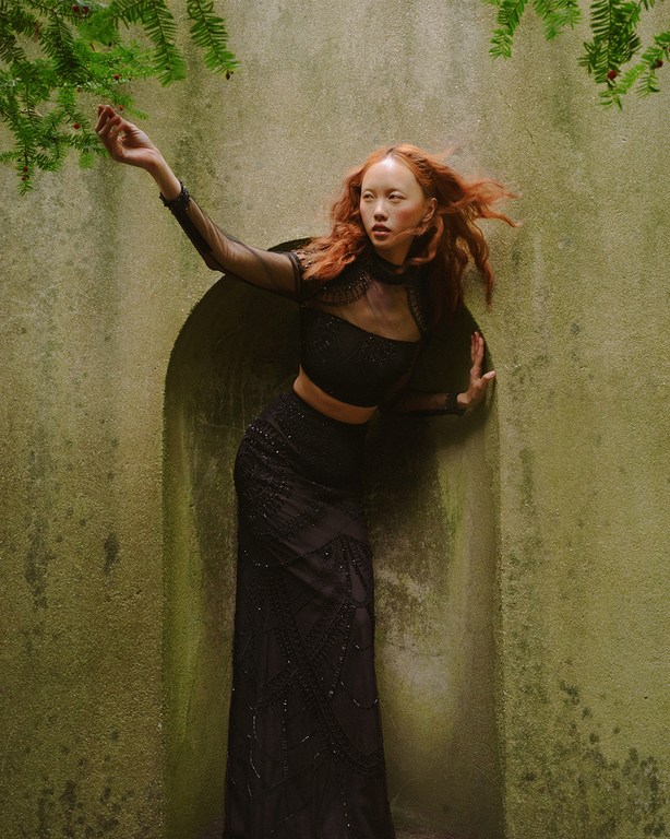 A red-haired woman in a black dress poses near a moss-covered wall and green foliage, lit by soft natural light in an outdoor setting.