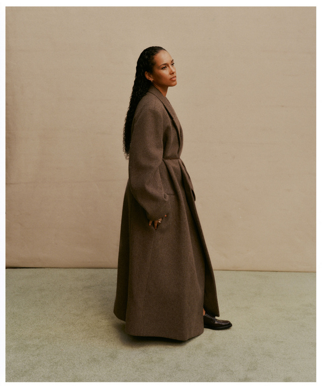 Alicia Keys in a brown wool coat, standing indoors against a neutral backdrop, showcasing fashion and celebrity style with a serene look.