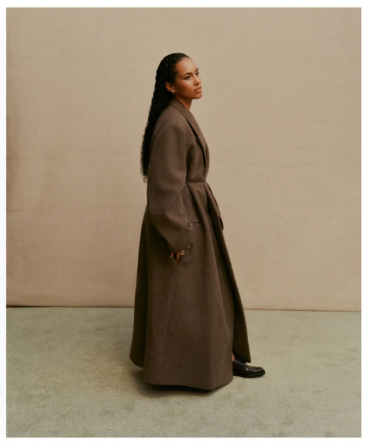 Alicia Keys in a brown wool coat, standing indoors against a neutral backdrop, showcasing fashion and celebrity style with a serene look.