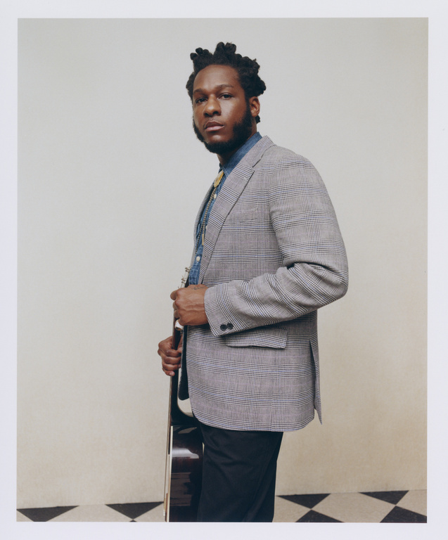 Portrait of an adult male musician wearing a suit jacket, dress shirt, and bolo tie, and holding a guitar inside a photo studio.