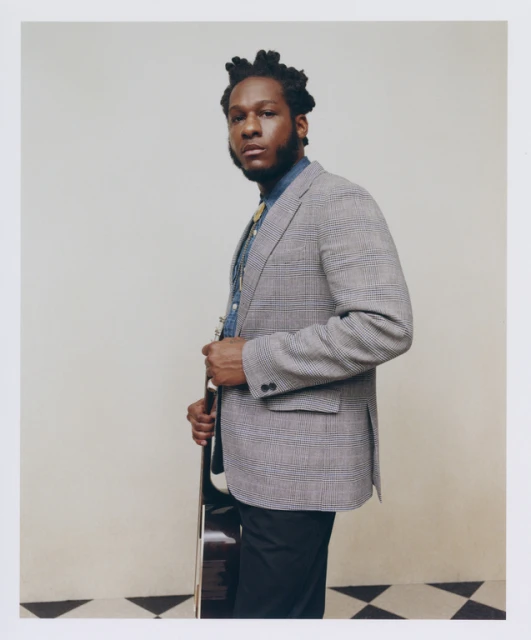 Portrait of an adult male musician wearing a suit jacket, dress shirt, and bolo tie, and holding a guitar inside a photo studio.