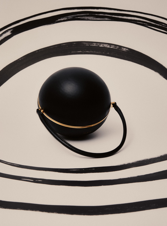 A black spherical purse with a gold accent handle is placed on a white surface with black painted semi-circular brushstrokes.