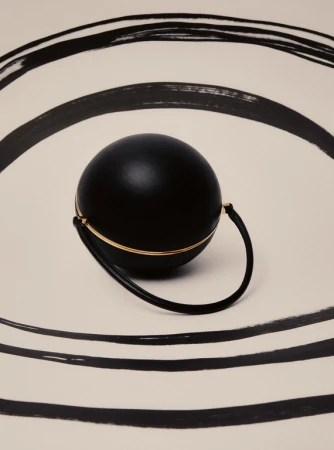 A black spherical purse with a gold accent handle is placed on a white surface with black painted semi-circular brushstrokes.