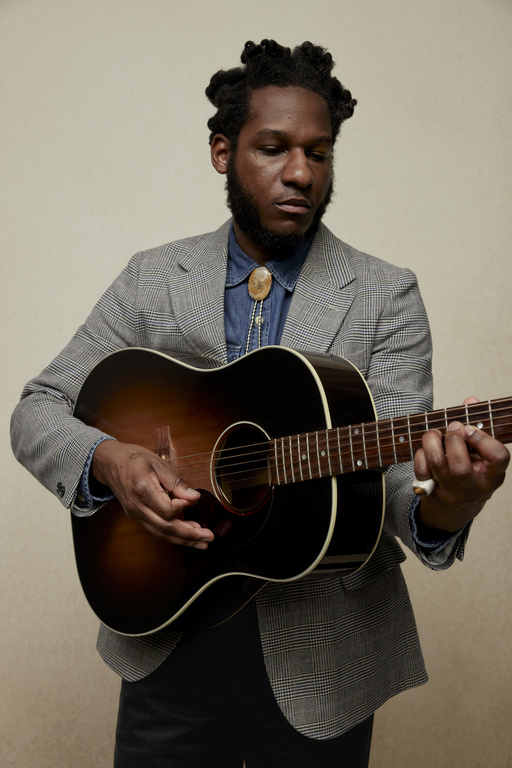 Man in tweed blazer plays acoustic guitar in a medium shot. He wears a bolo tie and denim shirt against a neutral backdrop.