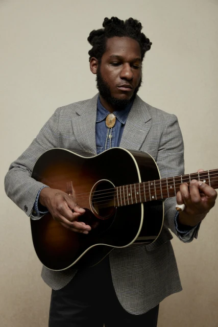 Man in tweed blazer plays acoustic guitar in a medium shot. He wears a bolo tie and denim shirt against a neutral backdrop.