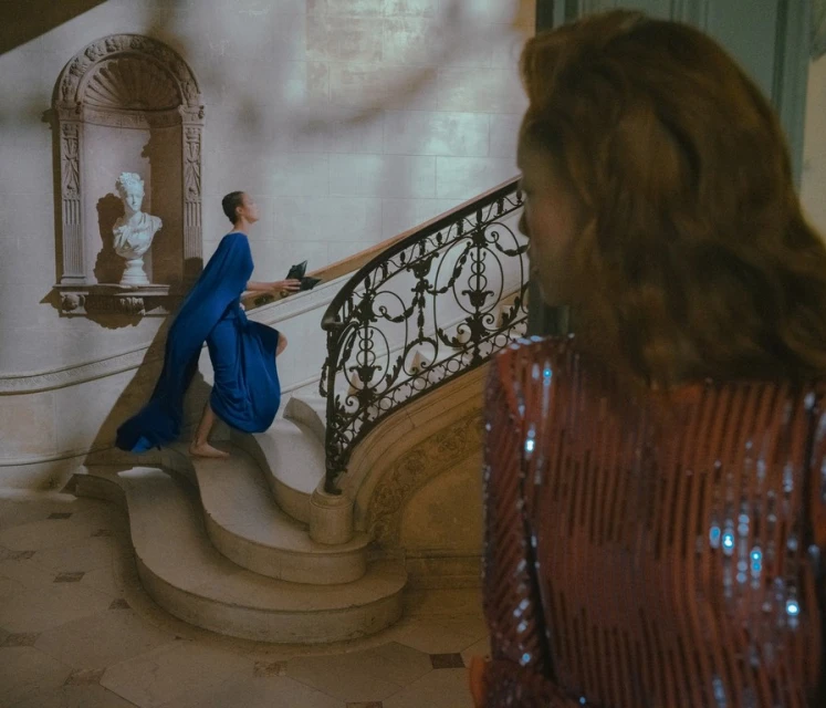 A woman in a blue gown climbs a staircase while another woman in a sequin dress stands in the foreground in an indoor setting.