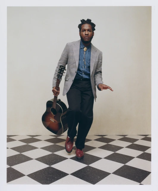 Adult male in gray blazer and denim shirt holding a guitar in front of a gray wall and checkered floor, fashion shot.