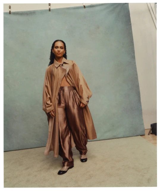 Woman in a stylish tan trench coat and silky pants poses in a studio setting against a blue backdrop, presenting a chic and modern fashion statement.