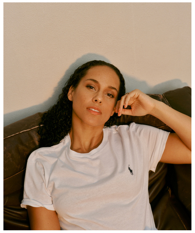 Attractive woman with long dark curly hair resting on a brown leather couch wearing a white short-sleeved T-shirt in a casual setting.