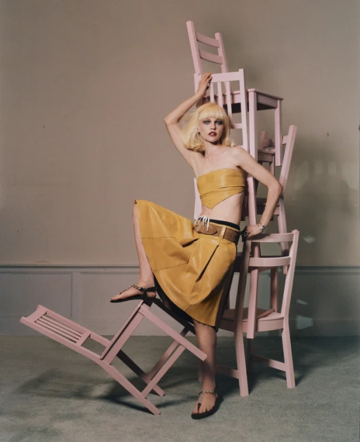 Fashion shot of a blonde model in a yellow outfit posing on pink folding chairs in a studio with soft lighting and neutral background.