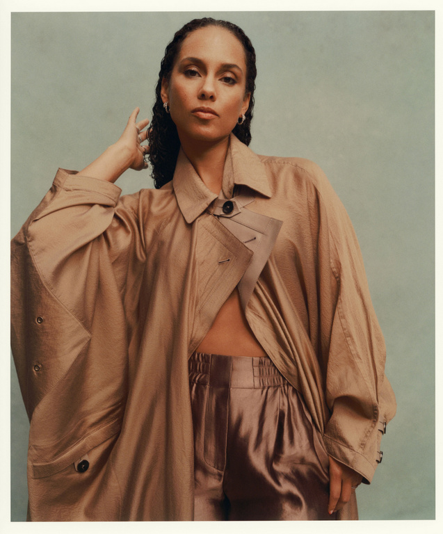 Portrait of a woman with wavy dark hair, wearing a loose tan trench coat and shiny bronze pants, against a pale greenish-gray background.