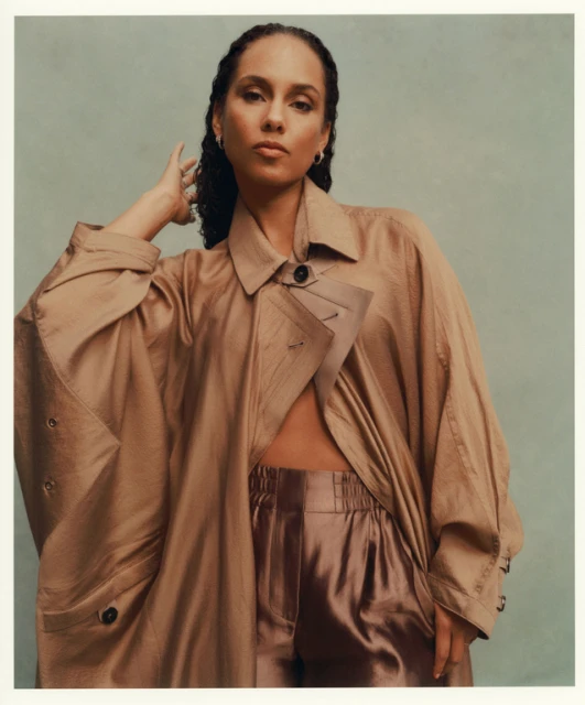Portrait of a woman with wavy dark hair, wearing a loose tan trench coat and shiny bronze pants, against a pale greenish-gray background.
