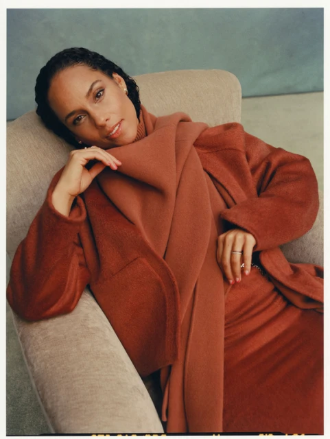 Close-up portrait of Alicia Keys in warm tones, wearing a cozy coat and scarf, sitting in a chair against a soft, neutral background.