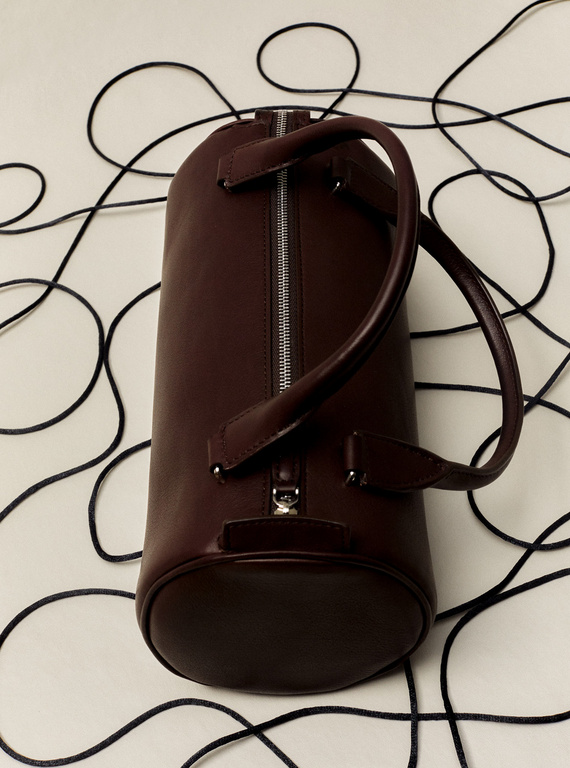 Cylindrical duffel bag in dark burgundy leather with metal zipper and handles on a white surface with dark, thin cords.