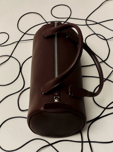 Cylindrical duffel bag in dark burgundy leather with metal zipper and handles on a white surface with dark, thin cords.