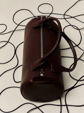 Cylindrical duffel bag in dark burgundy leather with metal zipper and handles on a white surface with dark, thin cords.