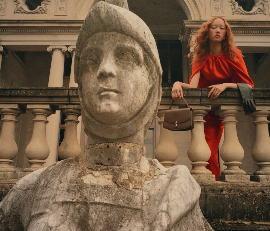 Woman in red dress poses on balcony with handbag near weathered stone sculpture and building with columns in background.
