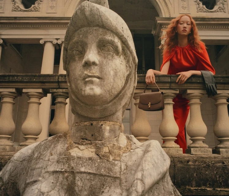 Woman in red dress poses on balcony with handbag near weathered stone sculpture and building with columns in background.