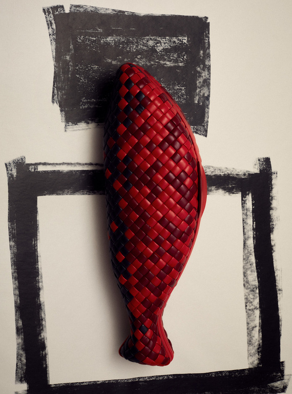 Still life of a red woven, fish-shaped object, centered against an off-white background with black rectangular borders brushed on it.