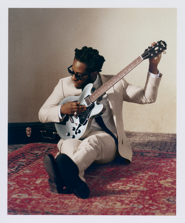 Adult male musician in a suit sits cross-legged on a patterned rug playing an electric guitar; a guitar case is in the background.