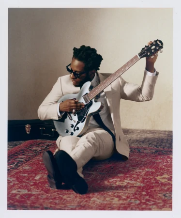 Adult male musician in a suit sits cross-legged on a patterned rug playing an electric guitar; a guitar case is in the background.