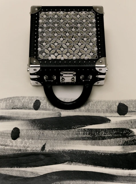 Stylish black trunk-style handbag with a silver geometric pattern on a white background with black and gray horizontal brush strokes.
