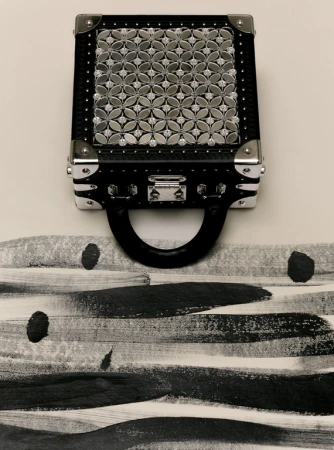 Stylish black trunk-style handbag with a silver geometric pattern on a white background with black and gray horizontal brush strokes.
