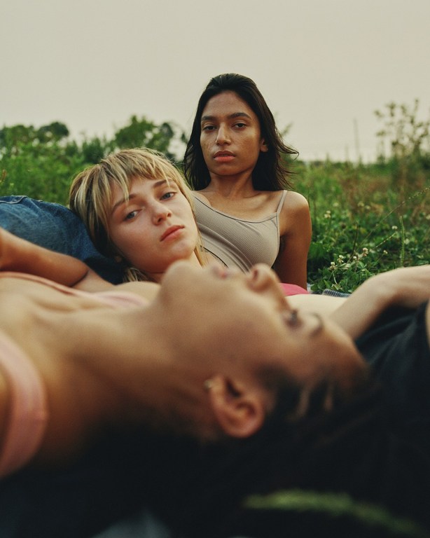 Three diverse young women lie outdoors on a blanket in an eye-level shot, wearing casual tank tops in a verdant field, with natural light.