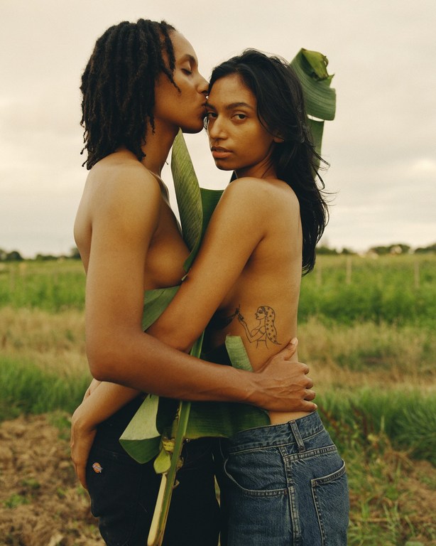 Two adults embrace outdoors in a field; one kisses the other's forehead, both partially concealed by a large, green leaf. 
