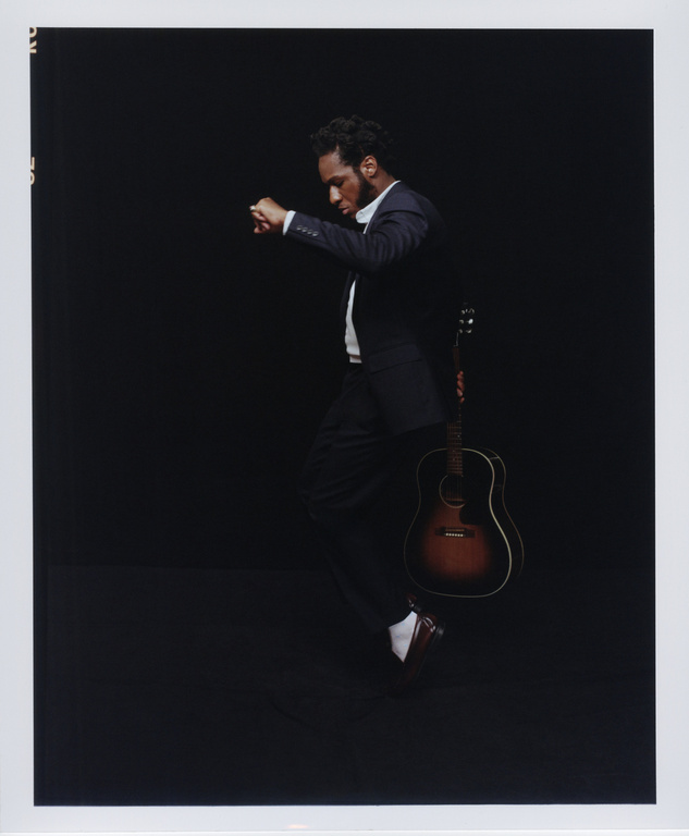 A man in a suit leans back with a guitar on a black background. He is in motion, dancing or walking with elegance.