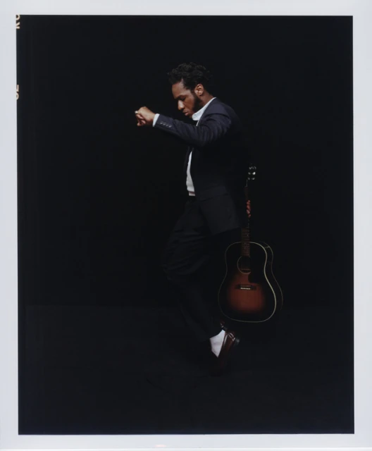 A man in a suit leans back with a guitar on a black background. He is in motion, dancing or walking with elegance.