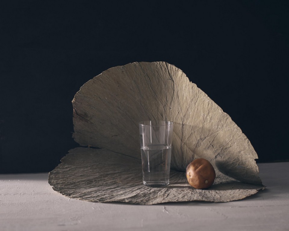 Still life photography features a glass of water, a potato, and a textured paper-like material on a white surface against a dark background.