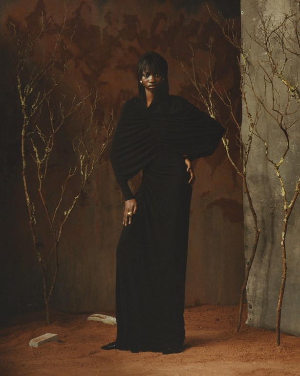 An elegant Black woman poses in a long black dress in an earthy-toned setting with bare tree branches, studio lighting, and a sophisticated look.
