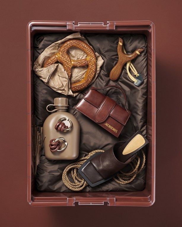 Still life shot of brown-themed objects including a brown leather purse, water bottle, pretzel, slingshot, rope, and heeled sandal inside of a brown bin.
