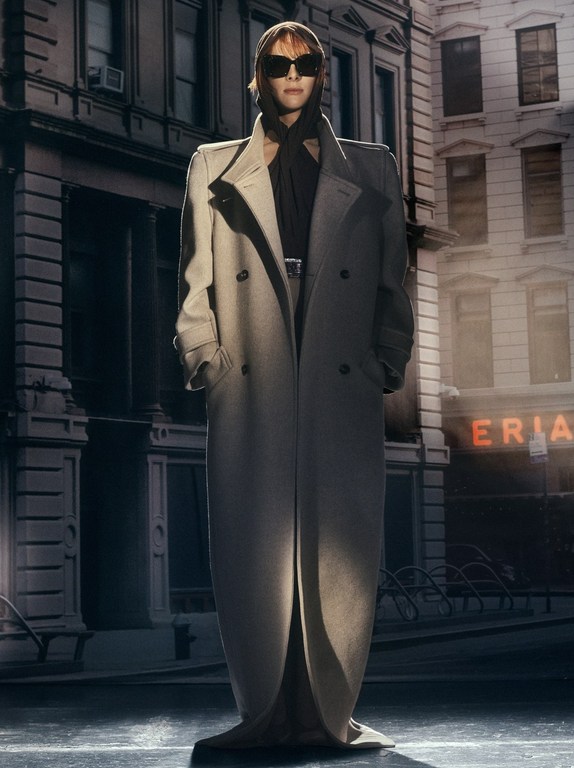 A woman stands in a beige overcoat, black headscarf, and sunglasses on an urban street with a building in the background.