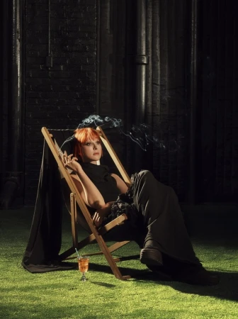 A woman with short red hair lounges in a wooden chair on faux grass, holding a cigarette with smoke, and a cocktail sits nearby.