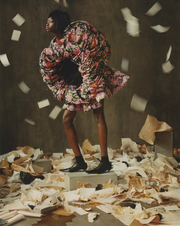 Fashion editorial shot of a Black model in a rose print, oversized dress standing on a concrete block surrounded by scattered papers.