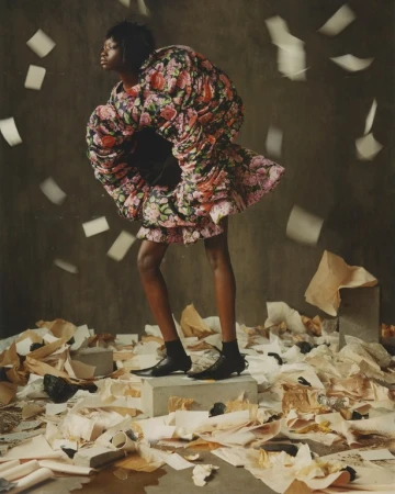 Fashion editorial shot of a Black model in a rose print, oversized dress standing on a concrete block surrounded by scattered papers.