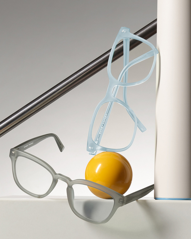 Still life studio shot of grey and blue eyeglass frames balanced on a yellow sphere against a white and grey background.
