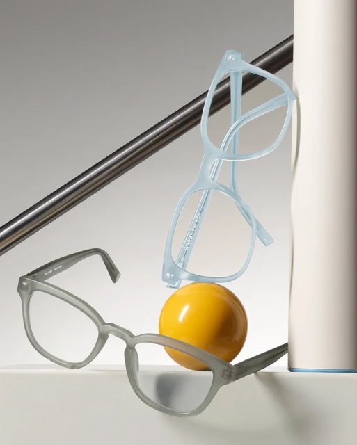 Still life studio shot of grey and blue eyeglass frames balanced on a yellow sphere against a white and grey background.