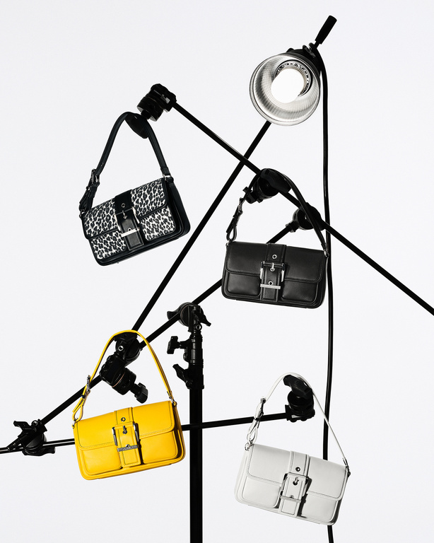 Four handbags of different colors and patterns, arranged on black metal stands against a bright white backdrop, illuminated by studio lighting.