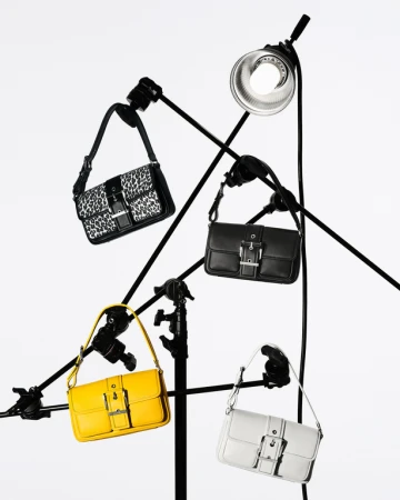 Four handbags of different colors and patterns, arranged on black metal stands against a bright white backdrop, illuminated by studio lighting.