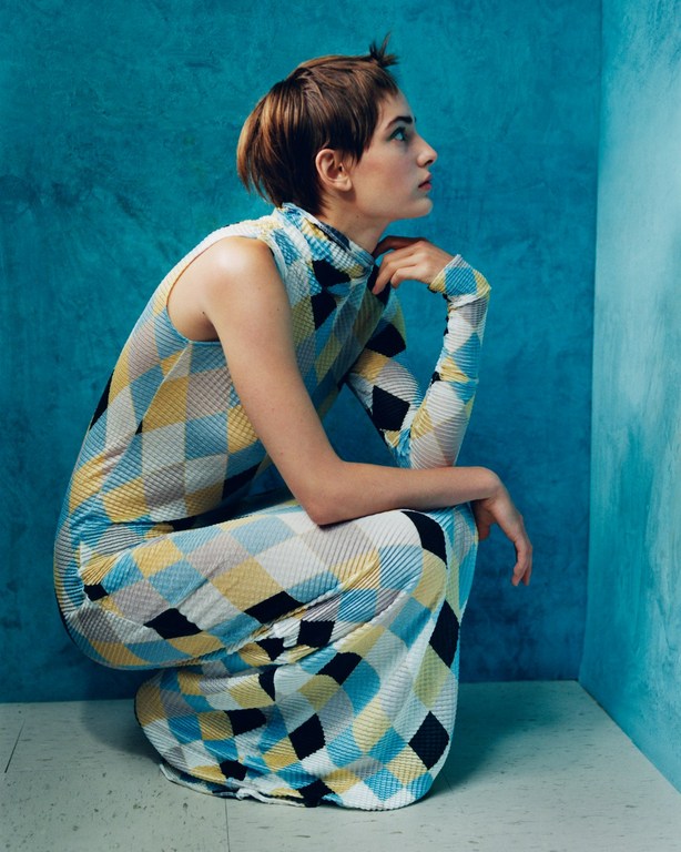 Fashion shot of a female model crouching against a blue corner wearing a patterned diamond dress with long sleeves and a high neckline.