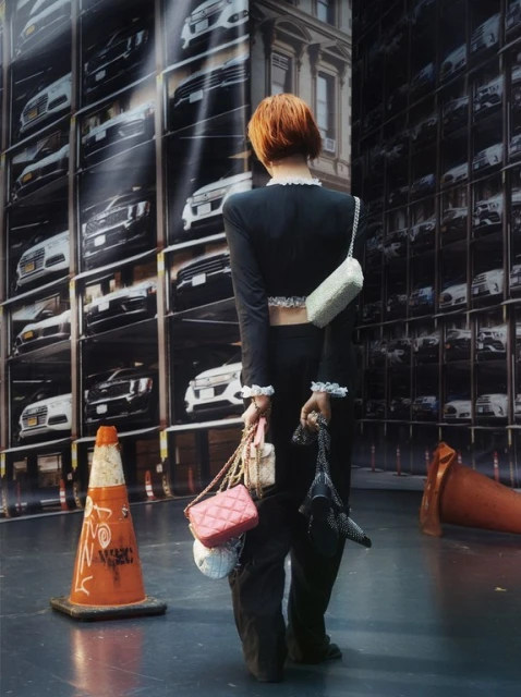 From a low angle, a stylish woman with red hair carries several handbags in front of a backdrop of a parking garage of cars.