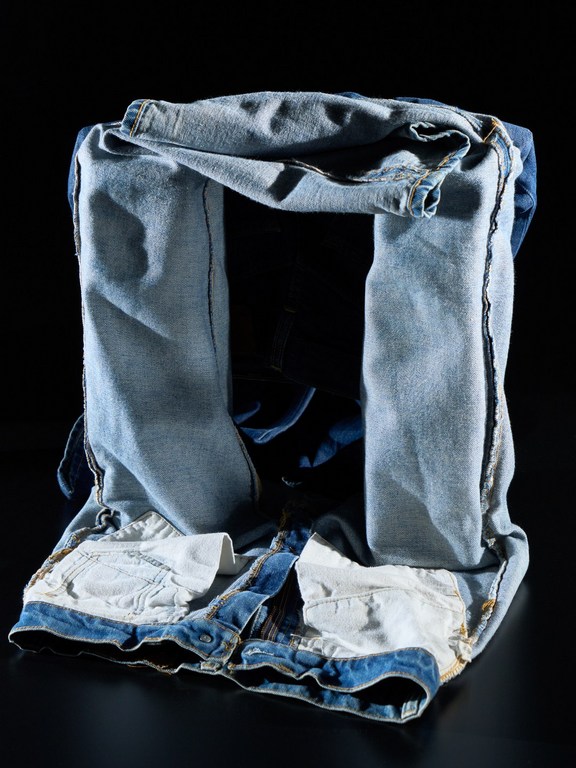 Close-up photo of stacked blue denim jeans in a square shape against a dark background, showcasing fabric details and textures.