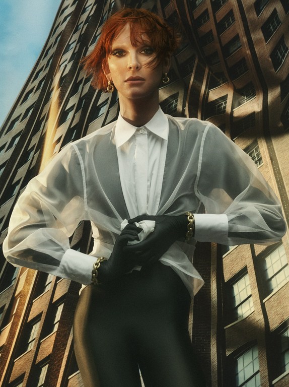 Stylish woman with red hair wearing a white shirt, gloves, and bracelet, posing against a building in an urban fashion portrait.
